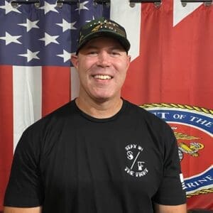 Smiling man in a black t-shirt and cap stands in front of American flags.